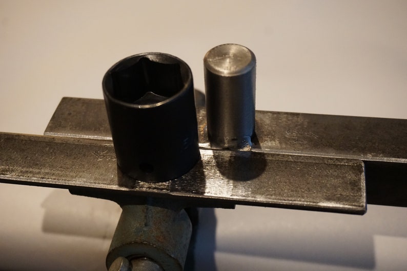 Op de afbeelding: Een close-up van twee metalen gereedschappen. Het gereedschap aan de linkerkant is een zwarte steeksleutel. Het gereedschap aan de rechterkant is een zilveren metalen staaf. Beide gereedschappen liggen op een metalen oppervlak.