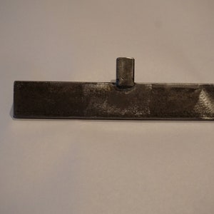Op de afbeelding: Een metalen gereedschap met een plat, rechthoekig blad en een klein, rechthoekig handvat. Het gereedschap is gemaakt van donkergrijs metaal en heeft een ruw, getextureerd oppervlak.