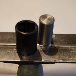 Op de afbeelding: Twee metalen voorwerpen, een zwart en een zilver, liggen op twee metalen staven. Het zwarte voorwerp is een dop en het zilveren voorwerp is een cilindrische dop.