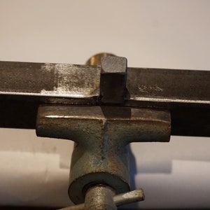 Op de afbeelding: Een metalen staaf is vastgeklemd in een bankschroef. De staaf heeft een vierkant stuk metaal dat aan de bovenkant is gelast.