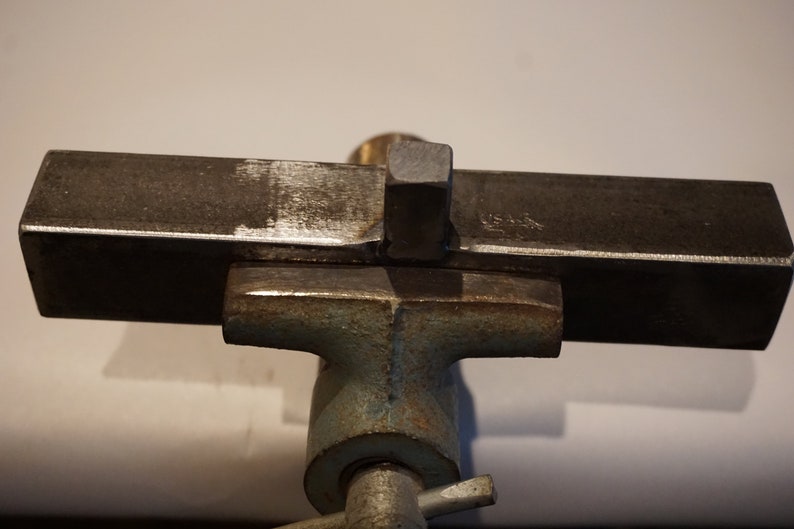 Op de afbeelding: Een metalen staaf is vastgeklemd in een bankschroef. De staaf heeft een vierkant stuk metaal dat aan de bovenkant is gelast.