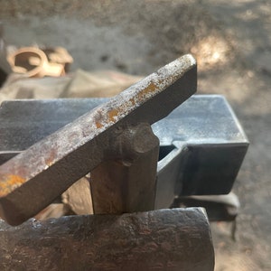 May include: A close-up of a metal anvil with a metal bar resting on top. The anvil is made of a dark metal and has a rough texture. The bar is made of a lighter metal and has a smooth texture.