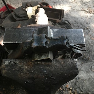 May include: A blacksmith's anvil with a forged metal dragon head sculpture. The dragon head is dark, detailed, and sits atop a rectangular piece of metal. Other metal pieces are in the background.