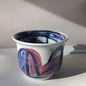 May include: A small white ceramic cup with blue and pink abstract designs. The cup has a rounded shape and a slightly flared rim.