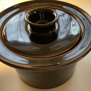 Puede incluir: Una fuente de cerámica marrón oscuro con tapa. La tapa tiene un pomo central y un borde ancho. La fuente es redonda y tiene un acabado brillante. El diseño es simple y funcional.