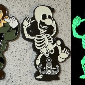 May include: Three enamel pins featuring a cartoon character. One pin shows the character in a green military outfit, covering their ears. Another pin depicts the character as a skeleton. The third pin glows in the dark.