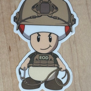 May include: A sticker depicting a cartoon character, similar to Toad, in military attire. The character is wearing a tan helmet and a vest with "EOD" on it. It holds a rope and a carabiner. The sticker has a white border and is on a wooden surface.