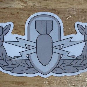 May include: A gray sticker featuring a military EOD (Explosive Ordnance Disposal) badge. The badge includes a bomb, lightning bolts, and a wreath design. The sticker is on a wooden surface.