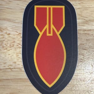 Puede incluir: Una pegatina con un diseño estilizado de una bomba roja con detalles amarillos y negros. La forma de la bomba está centrada en un fondo negro en forma de escudo.