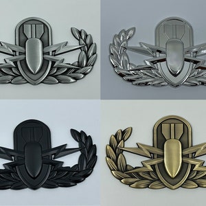 May include: Four metal badges in different finishes: silver, chrome, black, and bronze. Each badge features a central bomb design, flanked by lightning bolts and a laurel wreath. The badges are likely military or service emblems.