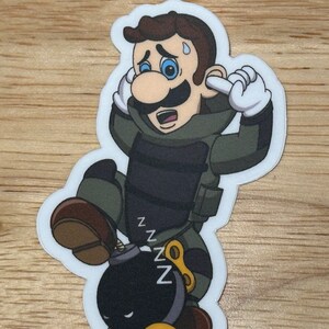 May include: A sticker featuring a cartoon character in a green and black outfit, covering their ears. The character is next to a black bomb with the word "zzz" above it. The sticker has a white border and is on a wooden surface.