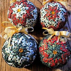 May include: Four handmade Christmas baubles. Each bauble is a sphere with a patchwork design in red, white, and green fabrics. They have gold ribbon bows and a small, gold-coloured gem at the centre.