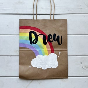 May include: A brown paper gift bag with a rainbow and a white cloud painted on it. The name "Drew" is written in black paint above the rainbow.