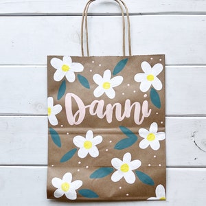 May include: A brown paper gift bag with a white daisy floral pattern and the name "Danni" written in pink paint.