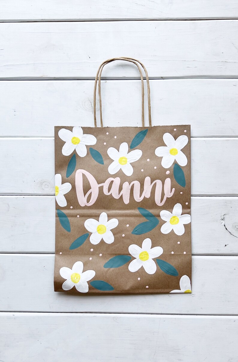 May include: A brown paper gift bag with a white daisy floral pattern and the name "Danni" written in pink paint.