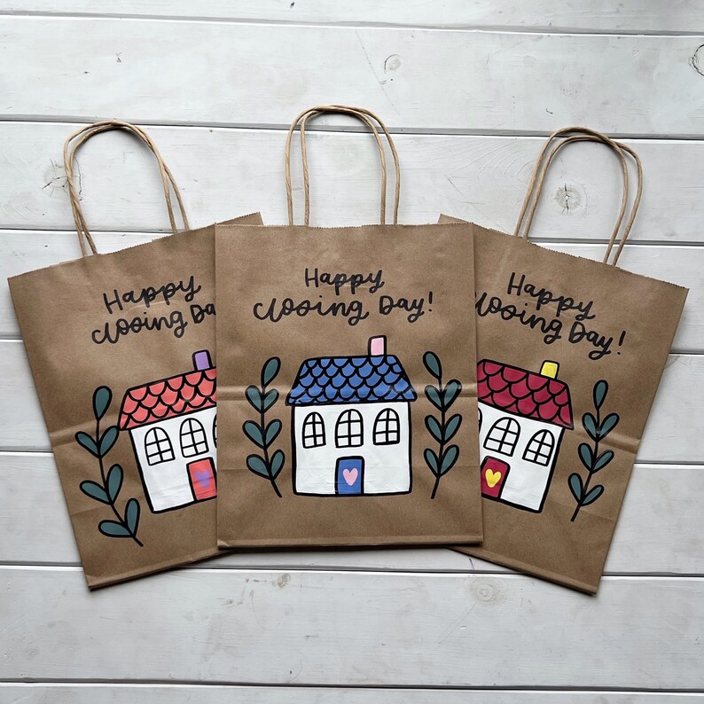 May include: Three brown paper gift bags with handles. Each bag has a hand-drawn house with a different colored roof and the text "Happy Closing Day!"