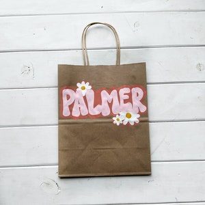 May include: A brown paper bag with a pink and white design. The bag has a handle and says "PALMER" in pink bubble letters with white daisies around the text.
