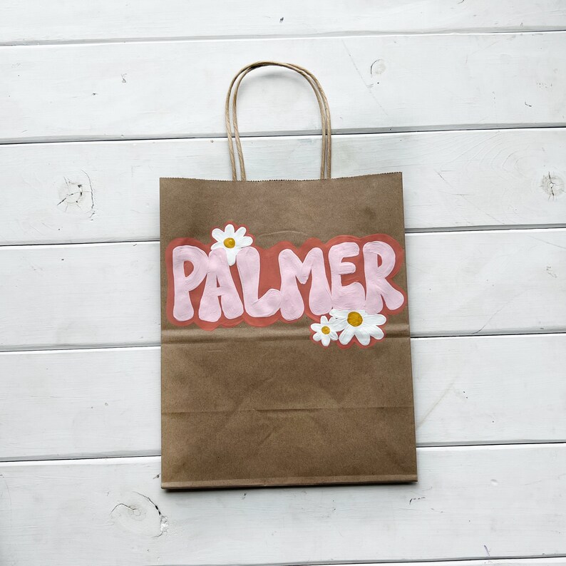 May include: A brown paper bag with a pink and white design. The bag has a handle and says "PALMER" in pink bubble letters with white daisies around the text.