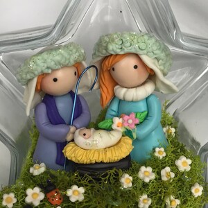 May include: A glass star-shaped ornament with a miniature nativity scene inside. Two figures, one in blue and one in purple, stand beside a manger with a baby inside. The scene is surrounded by green moss and white flowers.