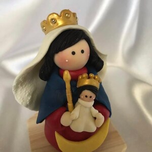 Könnte beinhalten: Eine handgefertigte Figur der Jungfrau Maria, die ein Jesuskind hält. Die Figur ist aus Polymer Clay gefertigt und in verschiedenen Farben bemalt, darunter Blau, Rot, Gold und Weiß. Die Jungfrau Maria trägt ein blaues Gewand mit einer goldenen Krone und einem weißen Schleier. Das Jesuskind trägt ein weißes Gewand mit einer goldenen Krone. Die Figur steht auf einem Holzsockel.