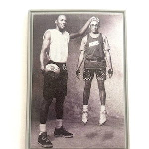 May include: Black and white photograph of two people, one holding a basketball. The person on the left is wearing a white vest top and shorts. The person on the right is wearing a short-sleeved shirt with the word "Nike" on it, shorts, and a headband. The photo is framed in a grey frame.