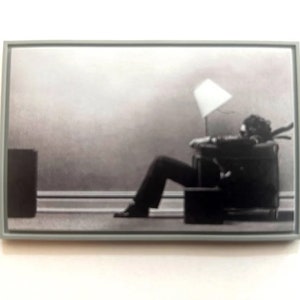 Könnte beinhalten: Gerahmte Schwarz-Weiß-Fotografie, die eine Person zeigt, die sich in einem Stuhl mit einer kleinen Lampe zurücklehnt. Die Person trägt dunkle Kleidung. Das Foto befindet sich in einem grauen Rahmen.