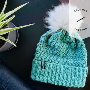 May include: A teal blue crochet beanie with a white pom pom. The beanie has a textured stitch pattern and a black tag that says "Hooked". The beanie is on a dark brown surface with a green plant in a gray pot.