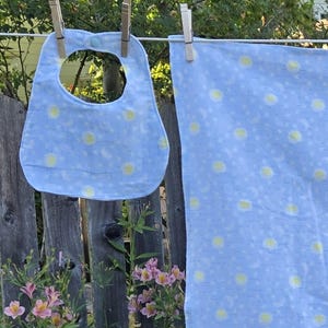 May include: A baby bib and a matching receiving blanket are hanging on a clothesline. Both items are light blue with a pattern of yellow suns and white crescent moons. The bib has a button closure.