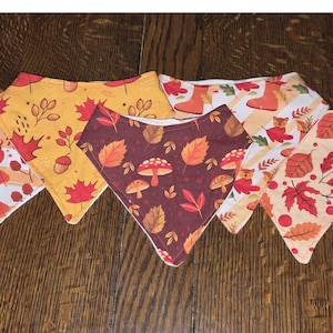 May include: A collection of six triangular baby bibs with autumn-themed patterns. The bibs feature designs of leaves, acorns, pumpkins, and mushrooms in shades of orange, yellow, and brown. Each bib has a white border.