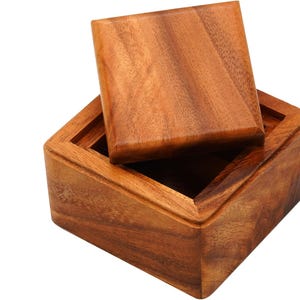 Op de afbeelding: Een houten kist met een iets geopende deksel, die de binnenkant laat zien. De kist is gemaakt van warm getint hout, met zichtbare nerfpatronen. Het vierkante deksel past netjes bovenop. De kist is waarschijnlijk voor opslag of weergave.