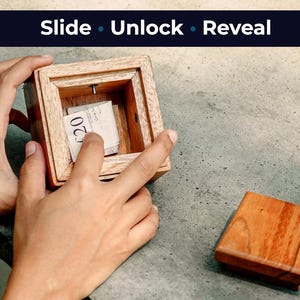 Op de afbeelding: Een houten puzzelbox met de woorden "Slide Unlock Reveal" bovenaan. De doos is open en onthult een stapel van twintig eurobiljetten. Het deksel van de doos ligt ernaast. De doos wordt door een persoon geopend.