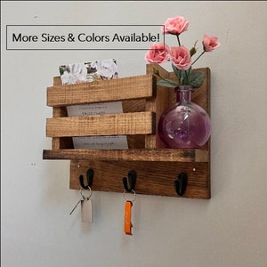 May include: Wooden wall mounted key holder with a shelf and a small mail slot. The shelf has three hooks for keys and a small vase with pink flowers. The text "More Sizes & Colors Available!" is printed on the top of the image.