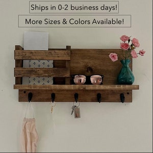 May include: A wooden wall-mounted key holder with a shelf and a mail slot. The key holder has five hooks and a shelf for storing items like sunglasses, keys, and mail. The shelf is made of rustic brown wood and has a natural wood finish. The key holder is mounted on a white wall.