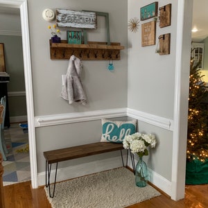 May include: A wooden entryway bench with black hairpin legs, a white fluffy rug, and a blue pillow with the word "hello" on it. A wooden shelf with hooks is mounted on the wall above the bench. The shelf has a gray coat hanging on one of the hooks. There are several wooden signs on the wall above the shelf, including one that says "gather".