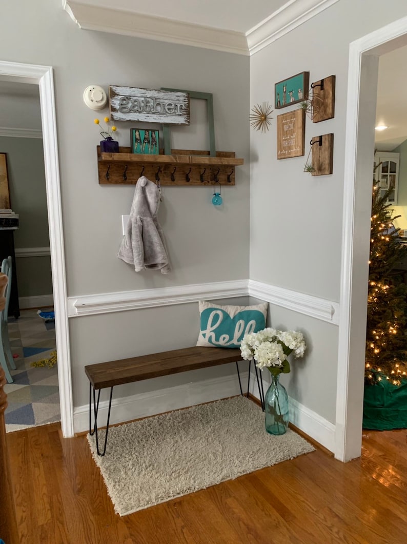 May include: A wooden entryway bench with black hairpin legs, a white fluffy rug, and a blue pillow with the word "hello" on it. A wooden shelf with hooks is mounted on the wall above the bench. The shelf has a gray coat hanging on one of the hooks. There are several wooden signs on the wall above the shelf, including one that says "gather".