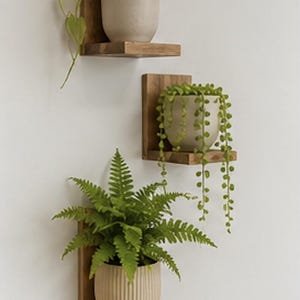 May include: Three individual L-shaped wooden wall shelves mounted vertically. Each shelf holds a potted plant: a trailing pothos in a smooth beige pot, a string of pearls in a smooth beige pot, and a fern in a vertically ribbed beige pot.