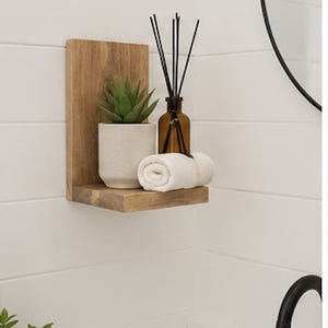 May include: A light brown wooden L-shaped wall shelf holding a small green succulent in a textured white pot, a brown glass bottle with black reed diffuser sticks, and a neatly rolled white hand towel, mounted on a white horizontal paneled wall.