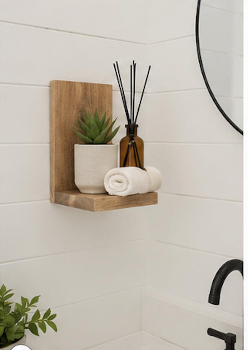 May include: A light brown wooden L-shaped wall shelf holding a small green succulent in a textured white pot, a brown glass bottle with black reed diffuser sticks, and a neatly rolled white hand towel, mounted on a white horizontal paneled wall.