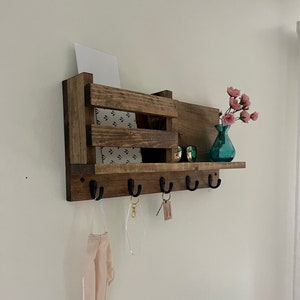 May include: A wall-mounted wooden organizer featuring a dark brown finish, a slatted storage compartment, and five black metal hooks. The shelf holds a white card, a pair of sunglasses, and a small teal glass vase with pink flowers.