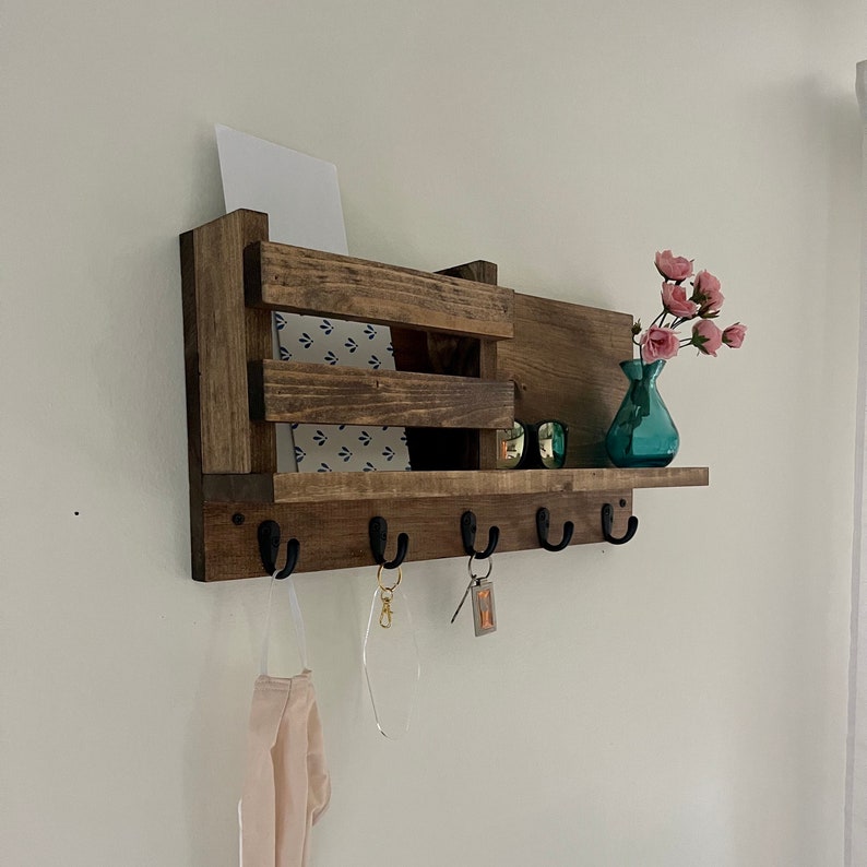 May include: A wall-mounted wooden organizer featuring a dark brown finish, a slatted storage compartment, and five black metal hooks. The shelf holds a white card, a pair of sunglasses, and a small teal glass vase with pink flowers.