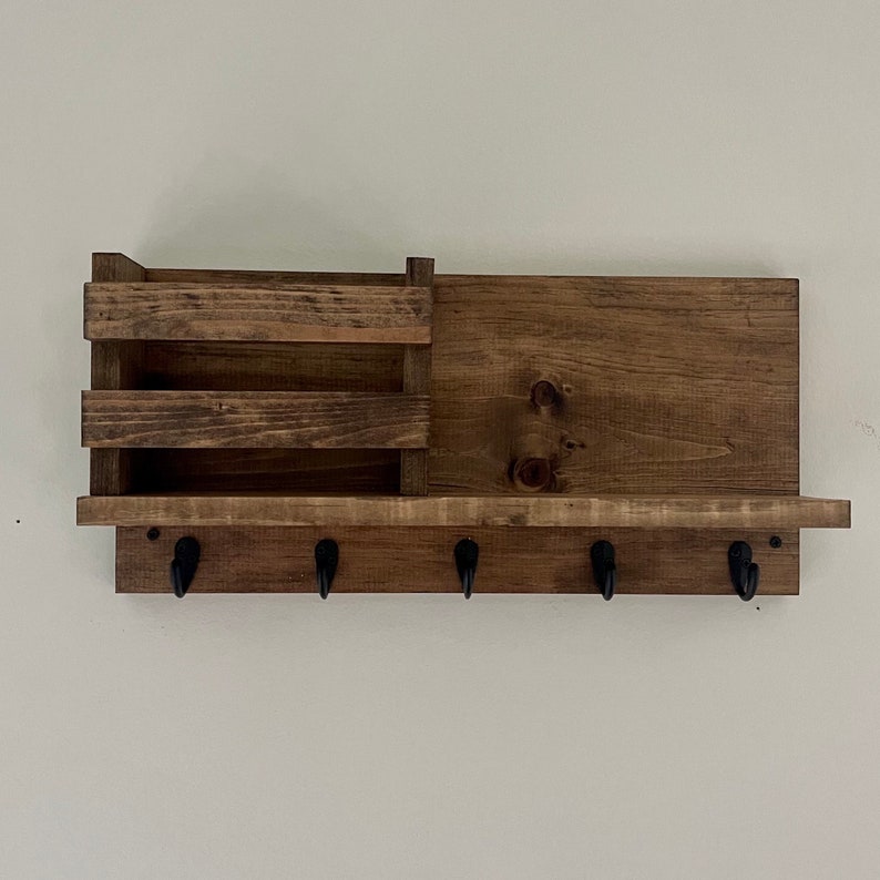 May include: A dark brown wooden wall organizer featuring a horizontal shelf with a built-in mail slot compartment on the left side. The unit includes five black metal hooks attached to the bottom edge for hanging items, showcasing a visible wood grain.