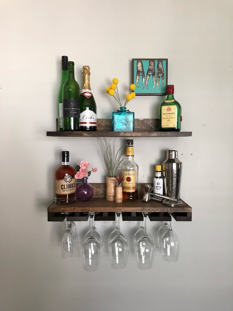 May include: A two-tiered, rustic wooden bar shelf with various bottles of liquor, wine glasses, and decorative items. The top shelf holds green and gold bottles, a small blue vase with yellow flowers, and a framed picture. The bottom shelf has whiskey, a cocktail shaker, and wine glasses.