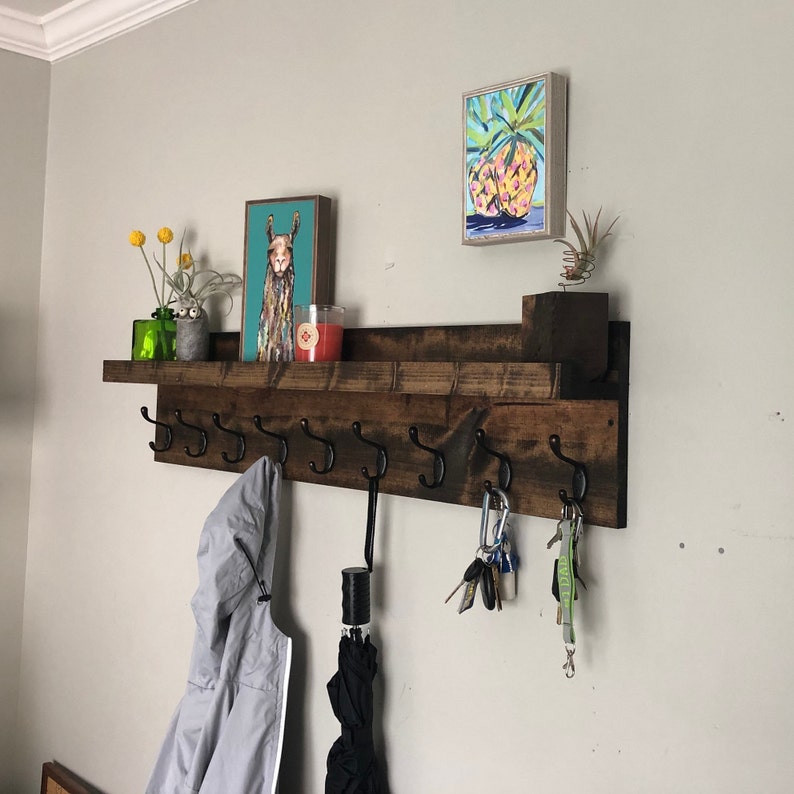 May include: A dark brown wooden shelf with metal hooks, a white shelf, and a llama print on the wall. There are keys, a green plant, and a candle on the shelf.