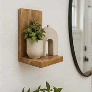 May include: A wall-mounted wooden shelf with a vertical back panel. On the shelf sits a small, light-colored ceramic pot containing a green leafy plant and a beige, arch-shaped decorative object with a small cylindrical top detail.