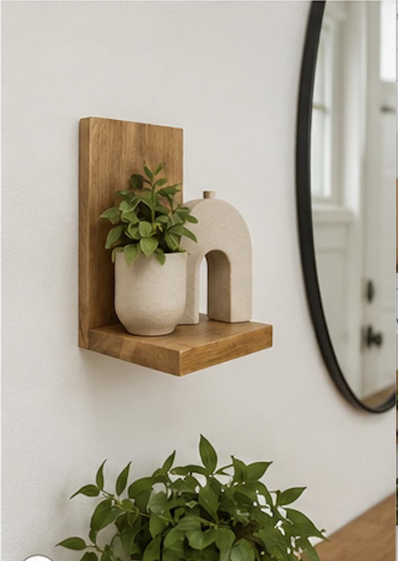 May include: A wall-mounted wooden shelf with a vertical back panel. On the shelf sits a small, light-colored ceramic pot containing a green leafy plant and a beige, arch-shaped decorative object with a small cylindrical top detail.