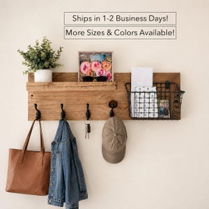 May include: A rectangular wooden wall organizer featuring a shelf, four black metal hooks, and a black wire storage basket. A potted green plant, a floral art print, a brown leather tote, a denim jacket, keys, and a tan baseball cap are displayed.
