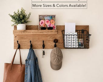 May include: A rectangular wooden wall organizer featuring a shelf, four black metal hooks, and a black wire storage basket. A potted green plant, a floral art print, a brown leather tote, a denim jacket, keys, and a tan baseball cap are displayed.
