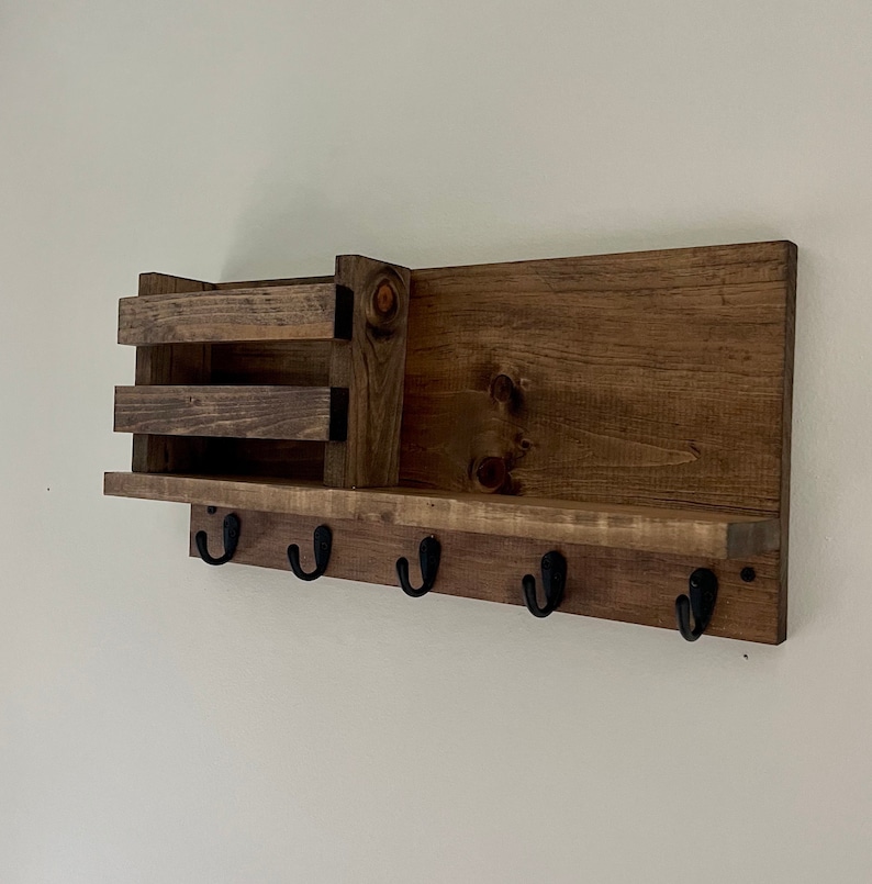 May include: A wall-mounted wooden organizer featuring a rectangular backboard with a dark brown finish. The unit includes a small storage bin on the left side and a horizontal shelf, with five black metal hooks attached to the bottom edge.
