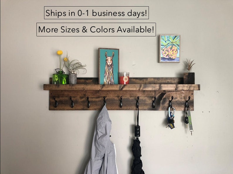 May include: A rustic wooden shelf with a coat rack underneath. The shelf is stained a dark brown and has a natural wood grain. The coat rack has six hooks and is made of black metal. There are various items hanging on the hooks, including a jacket, an umbrella, and keys. There are two small paintings on the wall above the shelf, one of a llama and one of a pineapple.