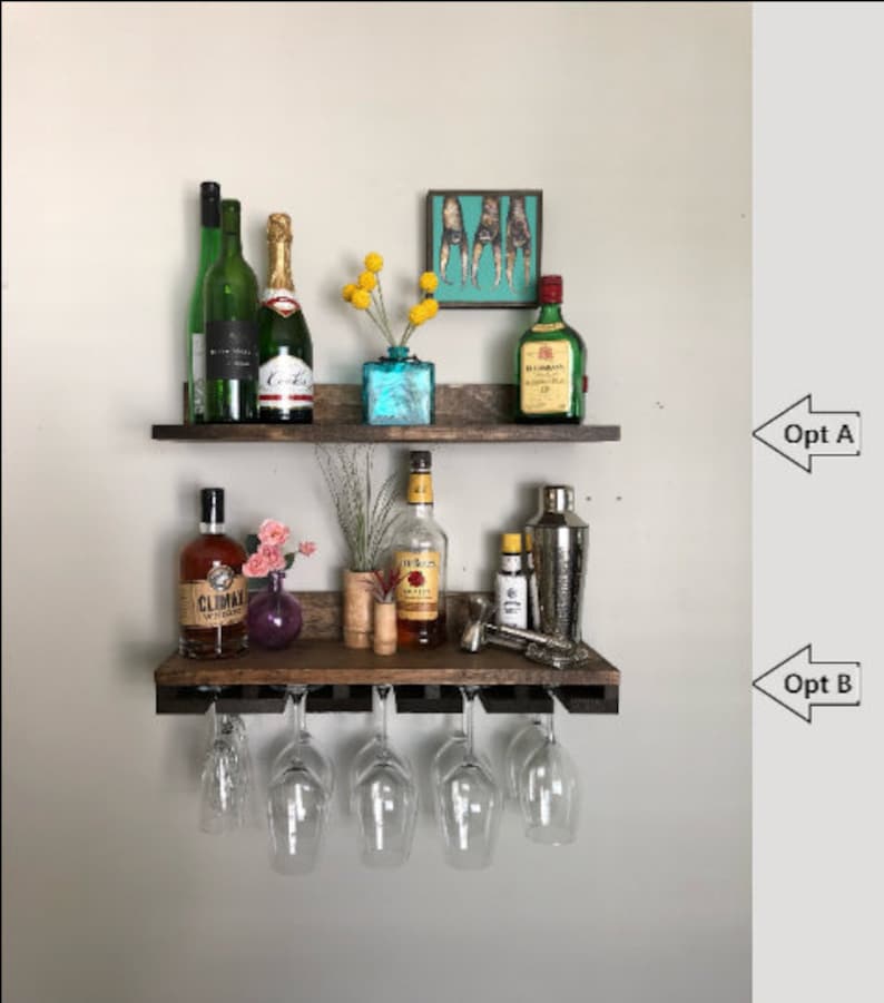 May include: A rustic, two-tiered wooden bar shelf with various bottles of liquor, champagne, and wine, along with cocktail glasses. The top shelf features a small painting and a blue vase with yellow flowers. The bottom shelf has wine glasses hanging below.